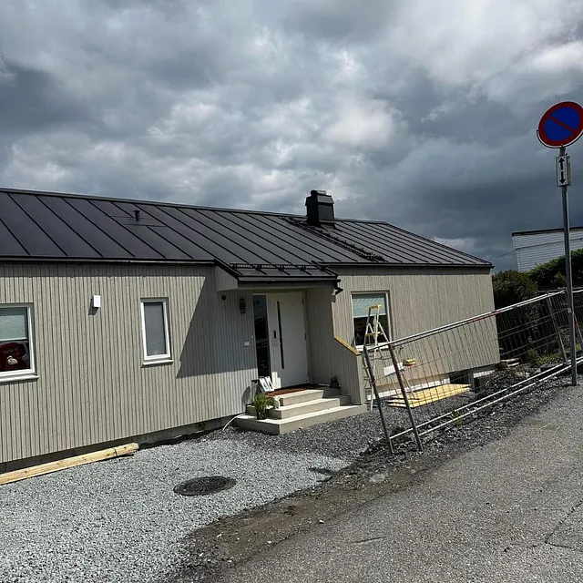 Grått hus med mørk blikktak og hvit inngangsdør, under en overskyet himmel.