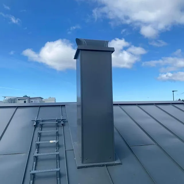 Stålpipe på tak med stige og blå himmel med lette skyer i bakgrunnen.