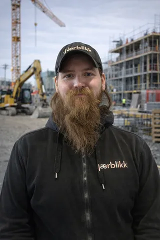 Mann med langt brunt skjegg og svart caps, på en byggeplass